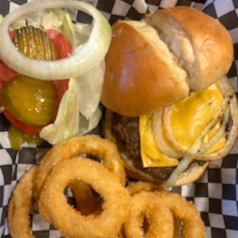 Cheeseburger with onion rings, lettuce, tomato, and pickles in a checkered paper-lined basket.
