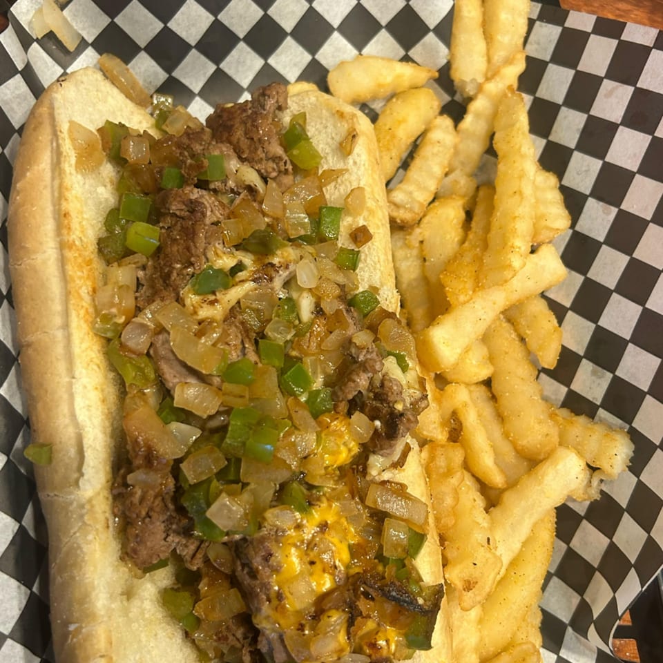 A cheesesteak sandwich with peppers and onions, served with crinkle-cut fries in a checkered basket.